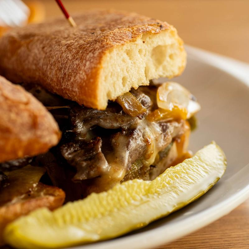 Steak sandwich on ciabatta with melted cheese, peppers, onions, and pickle served at 5800 North Cafe