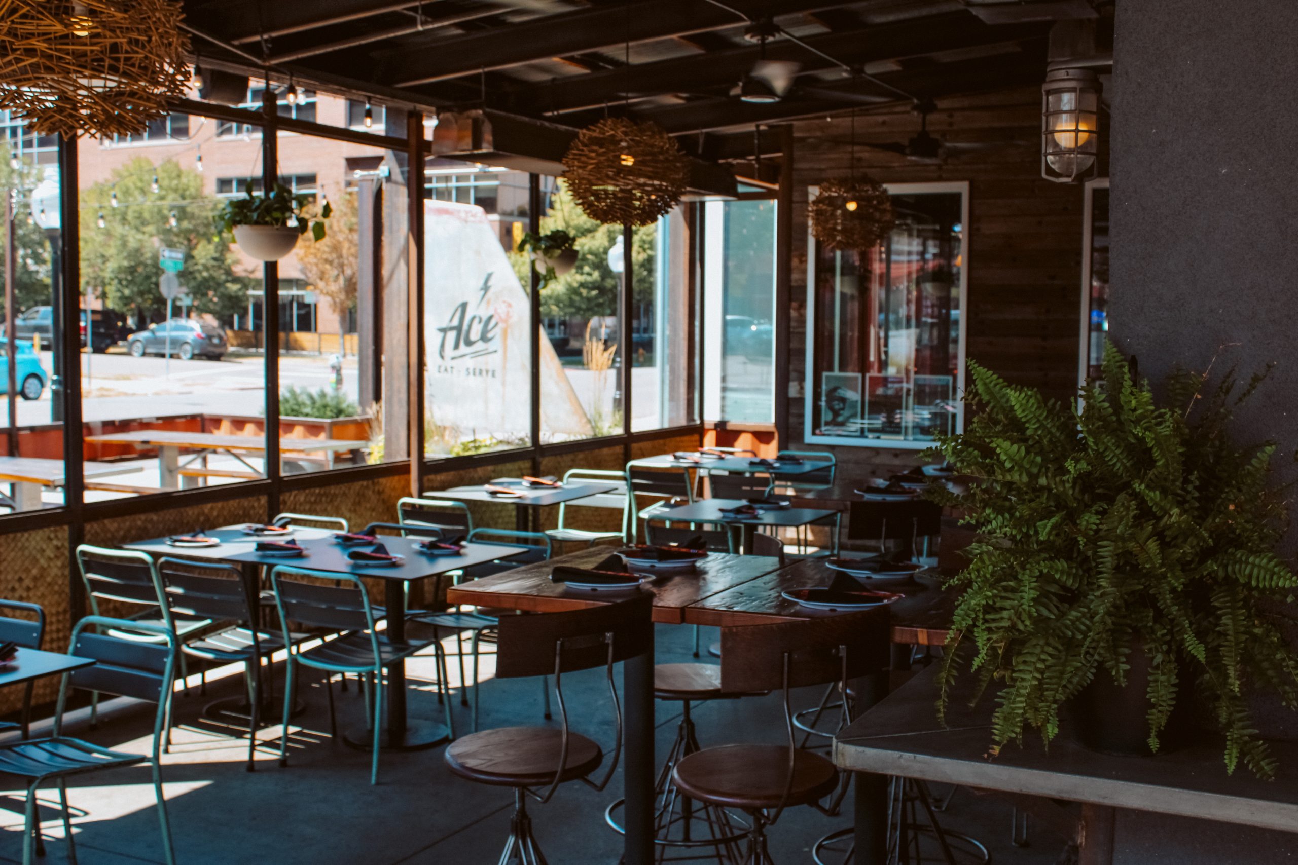 Covered patio at Ace Eat Serve with teal chairs, wooden tables, hanging lights, and greenery beside large street-facing windows.