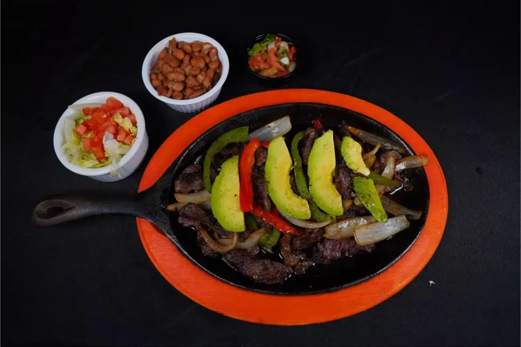 A cast-iron skillet filled with beef fajitas, grilled onions, bell peppers, and sliced avocado.